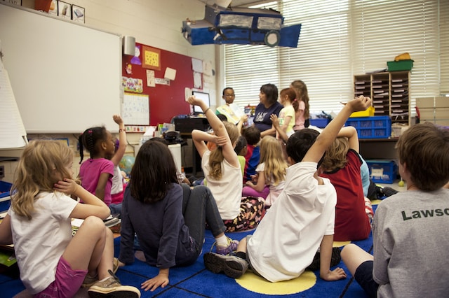 Nursery kids sitting around teacher with hands raised